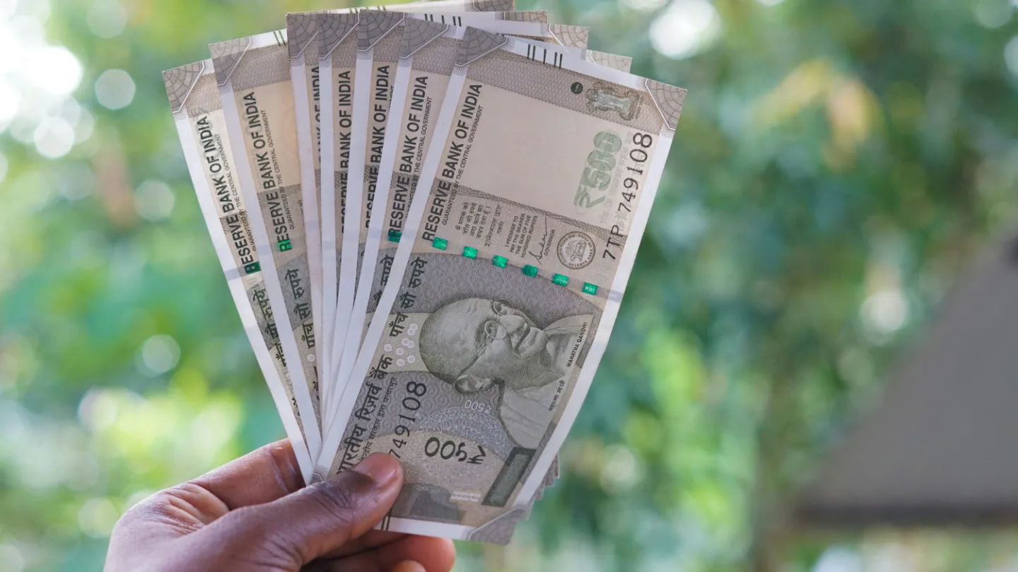 A hand holding a fan of seven ₹500 notes outdoors, against blurred green foliage.